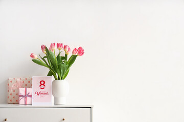 Vase with tulips, gifts and greeting card for Women's Day on chest of drawers near light wall