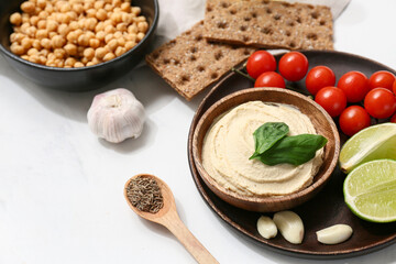 Bowl of tasty hummus with ingredients and crackers on white grunge background, closeup