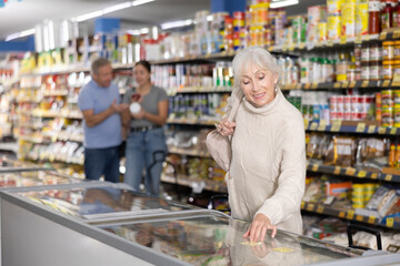 Attentive old woman purchaser choosing frozen product out of large stock in supermarket