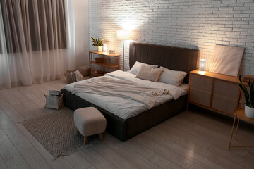 Stylish interior of bedroom with comfortable bed, soft pouf and glowing lamps at night