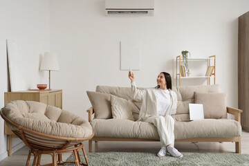 Young woman with laptop switching on air conditioner from sofa in living room