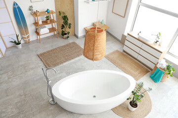 Interior of light bathroom with bathtub, big window, houseplants and surfboard