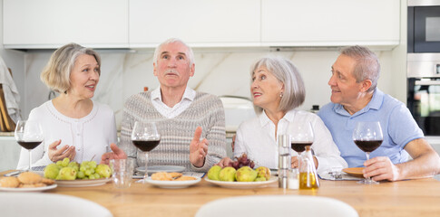 Four mature male and female friends are sitting at table, chatting sweetly, discussing and telling news, sharing plans. Senior women and men celebrates housewarming party with female relative.