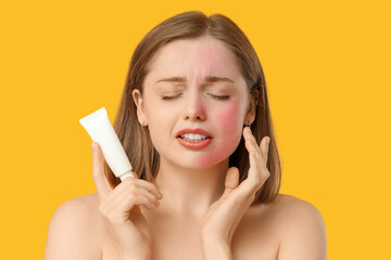 Suffering young woman with sunburned skin and sunscreen cream on yellow background