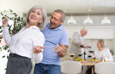 Cheerful mature friends dancing and drinking while enjoying home party on the room