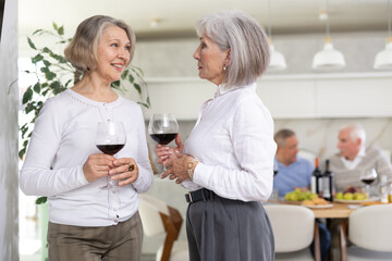 During friendly meeting, two elderly ladies with glass of wine in their hands stand at distance from table, communicate and chatting, smile, keep secrets.