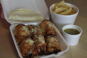 This image features a takeout box filled with grilled chicken thighs and tortillas served alongside...
