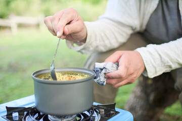 Unrecognizable person cooking a veggie lunch outdoors, rice with vegetables, using a portable gas stove, while camping. One hand holds the pot while the other stirs the food with a spoon.