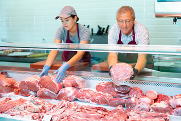 Focused skilled workers of butchery shop, elderly man and young woman, working together behind...