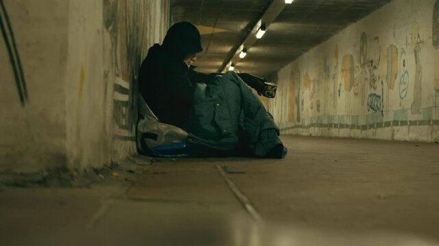 Hooded homeless man sits begging in cold dimly lit city urban subway