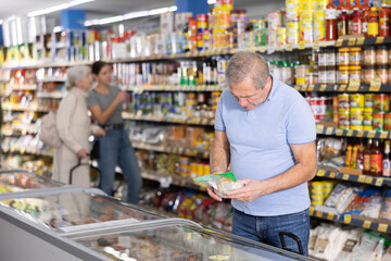 Senior man in casual jeans and t-shirt shopping for groceries in food store, looking at products in glass refrigerator with interest