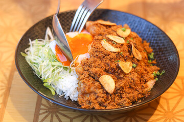 Close-up of crispy chicken with meat floss (abon), garlic, and melted soft-boiled egg cut by a fork and spoon over warm rice. A savory fusion of Indonesian and Japanese style dish.