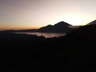 Stunning drone shot of Lake Batur at dawn, with mountain silhouettes reflecting on calm water under a colorful sky