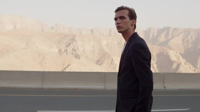 Confident young male model walks on road and looking camera. Handsome young guy model walking and turns around for camera. Male fashion model in a blue business suit looking back, on nature.