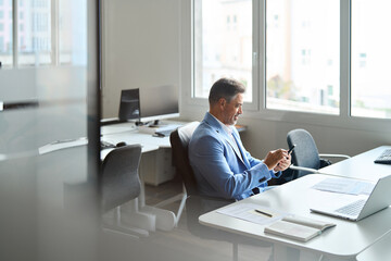 Happy busy middle aged business man investor wearing suit sitting in office using mobile phone. 50 years old smiling businessman professional banker holding smartphone at work. Authentic candid shot