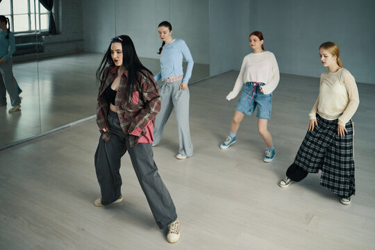 Group of young women perfecting dance routines in modern studio with mirrored wall and spacious floor building. Dancers wearing casual outfits creating diverse and collaborative atmosphere