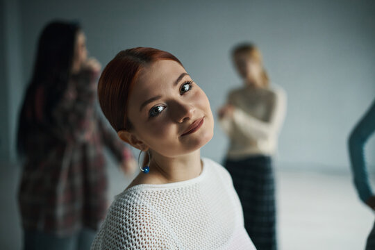 Portrait of young woman with reddish hair smiling at camera in room with blurred background of people. Capturing joyful expression and friendly atmosphere