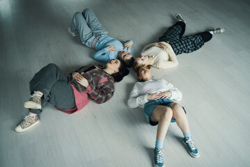 Four friends lying together on light wooden floor in casual attire, enjoying each other's company and sharing a relaxed moment. Captured from above with playful energy and connection