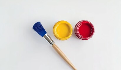  Minimalist white background with three paint jars in red, blue, and yellow next to one brush. Studio shot of an art setup for painting on a solid white backdrop.