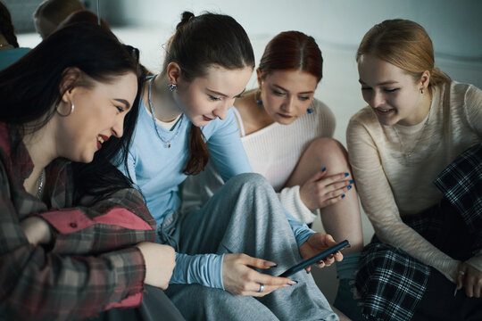 Group of young women sitting closely together, leaning in to share and view something on a smartphone while expressing collective amusement in cozy indoor setting