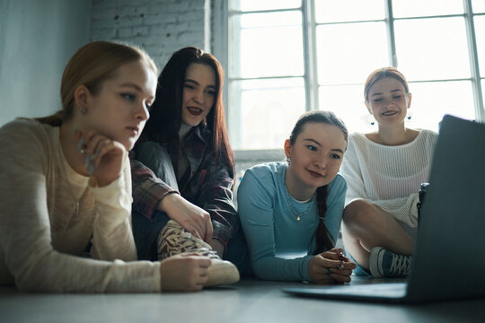 Group of teen girls gathering around laptop in bright room with large window and relaxed atmosphere, enjoying time together