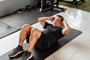 Southeast Asian man in his 50s performing abdominal crunches on a black mat during core workout at...