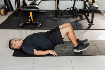 Southeast Asian man in his 50s performs glute bridge exercise on mat to activate glutes and core muscles during workout.