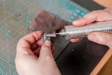 Hands of jeweler are using a rotary tool to grind a small silver pendant, on a green cutting mat.