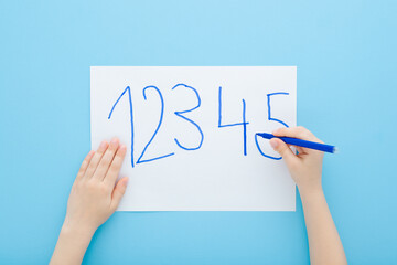Fototapeta premium Little child boy hand holding color pen and writing different numbers on white paper on light blue table background. Pastel color. Doing school exercise. Closeup. Point of view shot. Top down view.