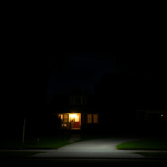 A dramatic nighttime scene of a power outage, where a single home remains illuminated thanks to its battery backup system. The surrounding houses are in darkness, with the glow of the powered home