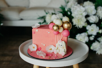 A cake for a child's birthday celebration lies on a round table, decorated with a decorative bunny toy holding inflatable balloons, the cake is pink in color