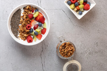 Top view of breakfast with white bowl of kefir, fruits, granola and chia seeds, wood and metal spoon, open jar with granola and square bowl with fruit on light stone background. 