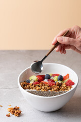 Female hand with spoon over white bowl with kefir, strawberries, kiwi, raspberries, blueberries, granola and chia. Peach background and gray stone surface. Healthy nutrition concept