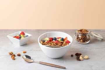 White ceramic bowl filled with kefir, granola, chia seeds and fresh berries, accompanied by a small square bowl with fruit and a glass jar with granola on a stone surface. 
