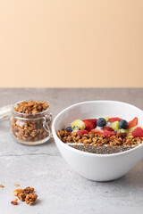Healthy breakfast bowl with kefir, chia seeds, granola and fruits on a light stone surface. A small jar of granola and some scattered granola pieces add texture to the scene.  