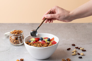 Front view of healthy breakfast with white bowl of kefir, strawberries, blueberries, raspberries, kiwi, granola and chia seeds. Female hand holding spoon over grey stone surface. 