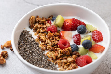 Top view of white bowl with kefir, strawberries, blueberries, raspberries, kiwi, crunchy granola and chia seeds. Light neutral grey background. Concept of balanced nutrition and healthy lifestyle
