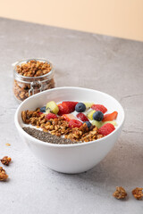 White bowl with kefir, strawberries, blueberries, raspberries, kiwi, granola and chia seeds on light stone surface. Granola jar in the background. Concept of healthy and natural breakfast