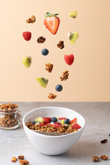 White bowl with fruits such as strawberries, raspberries, kiwi and blueberries, kefir, chia seeds and granola, with some fruits and granola suspended in the air. 