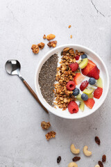 Vertical top-down shot of a white bowl with kefir, fresh fruits, granola and chia seeds on grey surface. On the left, a metal spoon; at the bottom, nuts. Concept of healthy breakfast and real food