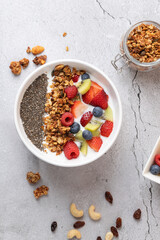 Vertical top view of a bowl with kefir, fruits, chia and granola on grey stone background. Carefully arranged composition with scattered nuts and two visible containers. 