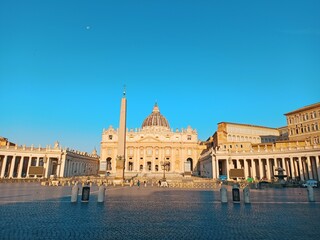 Obraz premium Stunning view of St. Peter’s Basilica with the Vatican Obelisk under a clear blue sky at sunrise. A symbol of Rome’s architectural and religious grandeur.