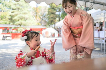 On a sunny spring day at a shrine, a Japanese woman in her thirties and her 3-year-old daughter in kimonos celebrate Shichi-Go-San. They draw omikuji, sharing a joyful moment in a peaceful setting.
