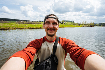 A cheerful, smiling man captures a delightful selfie by a stunning scenic lake while enjoying the beautiful partly cloudy skies. This picture represents joy and adventure during his vacation