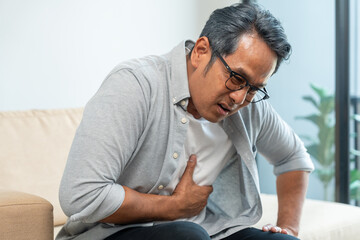 Fototapeta premium Old man senior he felt a pain in his chest held his hand and squeezed it. Mature Heart attack that needed treatment. Sitting on sofa couch in living room at house