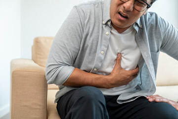 Fototapeta premium Old man senior he felt a pain in his chest held his hand and squeezed it. Mature Heart attack that needed treatment. Sitting on sofa couch in living room at house