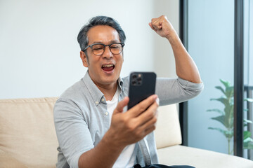 Handsome asian mature old man using smartphone on sofa in living room at home. Happy Portrait of cheerful smiling senior asian man holding cell phone. Mature People and lifestyle