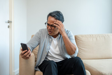 Depressed Asian mature old man using smartphone serious thinking on sofa in living room. Portrait stress of senior asian man holding cell phone. Mature People and lifestyle