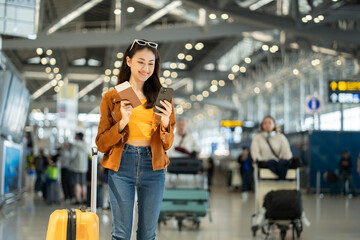 Happy young asian woman traveler using smartphone and holding boarding pass while standing at the airport terminal ready for vacation trip, smiling female tourist with confidently travel insurance