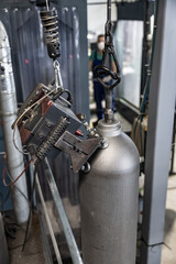 Close-up of a metallic gas cylinder being tested or inspected by an automated clamp device, suspended by a hook in an industrial environment